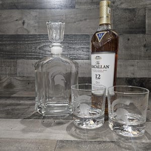 May include: A clear glass decanter with a etched design of a Spartan helmet, two clear glass tumblers with etched Spartan helmets, and a bottle of The Macallan 12 Single Malt Scotch Whisky.