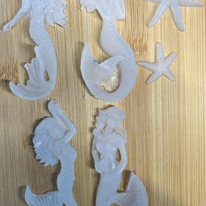 May include: Five translucent mermaid and starfish figurines. The mermaids have flowing hair and detailed tails. Two starfish are also visible. The figurines are arranged on a light brown wooden surface.