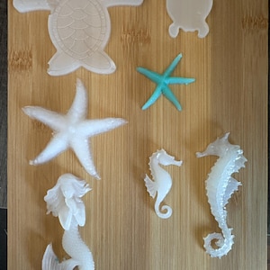 May include: A collection of sea-themed molds, including two turtles, two seahorses, a starfish, a mermaid, and a turquoise starfish. The molds are white and arranged on a wooden surface. Ideal for crafting projects.