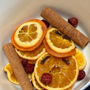 May include: A white bowl containing dried orange slices, cinnamon sticks, cloves, and red berries. The orange slices are stacked, and the cinnamon sticks are a rich brown. The cloves are small and dark, and the berries are a deep red.