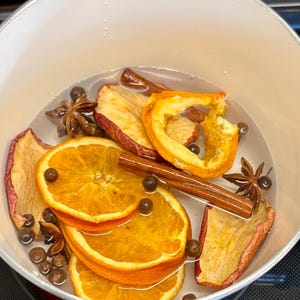 Może przedstawiać: Biały garnek z czerwonym obramowaniem wypełniony wodą, plasterkami pomarańczy, plasterkami jabłka, laskami cynamonu, anyżem gwiazdkowatym i ziarnami pieprzu. Składniki są zanurzone w wodzie, tworząc ciepłą, zachęcającą scenę.