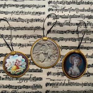 May include: Three round, gold-framed, miniature portraits with black ribbon hangers. The portraits feature a couple in a garden, a woman in a white dress, and a woman in a blue dress.