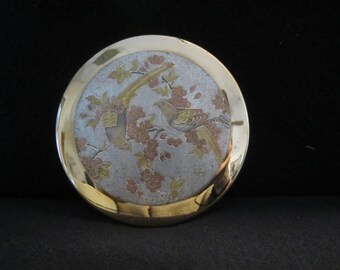 Könnte beinhalten: Runder, vintage Metall-Kompaktbehälter mit goldfarbenem Rand und detailliertem Vogel- und Blumendesign. Das zentrale Bild zeigt einen Vogel, der auf einem Ast mit rosa und goldenen Blüten vor silbernem Hintergrund sitzt. Ein dekorativer Artikel.