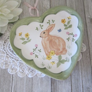 Peut inclure: Un plat de service en céramique en forme de cœur avec une bordure verte et un motif peint d'un lapin brun entouré de fleurs jaunes et roses.