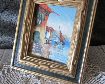 May include: A small framed painting of a seascape with a building in the foreground. The painting is in a gold and green frame.