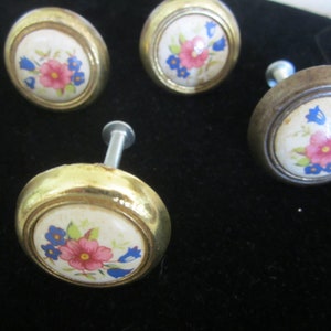 May include: Four gold-toned cabinet knobs with a white ceramic base and floral designs. The flowers are pink, blue, and yellow.