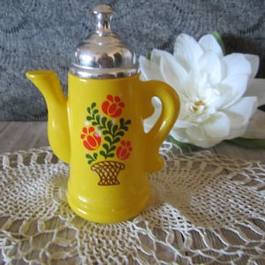 Puede incluir: Tetera de cerámica amarilla con tapa plateada y diseño floral. La tetera tiene asa y pico. El diseño floral presenta flores rojas, naranjas y verdes en una cesta.