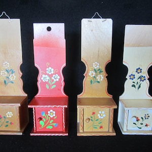 May include: Four wooden wall mounted storage boxes with painted floral designs. The boxes are painted in red, yellow, and white with a white trim. Each box has a small drawer with a knob.