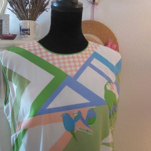 May include: A sleeveless, patterned dress featuring a geometric design in green, blue, and peach, with bird illustrations. The neckline has a checkered pattern. The dress is displayed on a black mannequin.