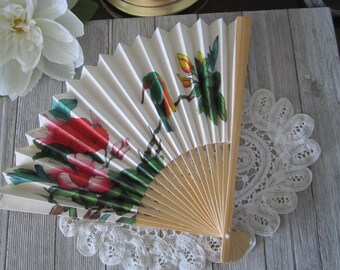 Puede incluir: Un abanico plegable blanco con un mango de bambú. El abanico presenta un diseño floral colorido con flores rojas, rosas y verdes y un pájaro verde.