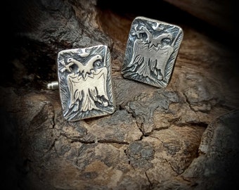 May include: A pair of silver cufflinks with a detailed design of two eagles facing each other. The cufflinks are on a rustic wooden background.