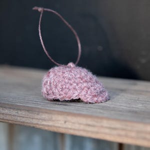 May include: A small, dusty rose-colored knitted cap with a string loop for hanging. The cap is resting on a wooden surface, with a dark background. The cap appears to be handmade.