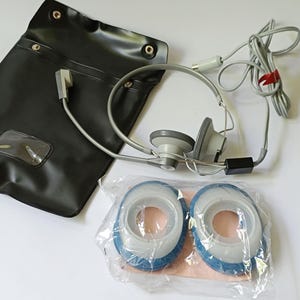 May include: A vintage grey headset with a black carry case and a pair of replacement ear cushions. The headset has a microphone and a coiled grey cable. The ear cushions are blue and white.