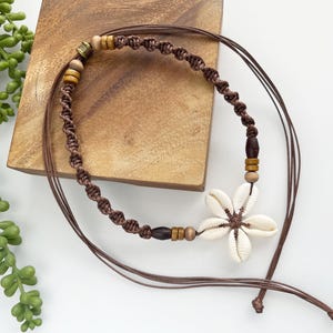 May include: A brown macrame necklace featuring a white seashell flower pendant. The necklace has wooden beads and a knotted design. The necklace is displayed on a wooden block.