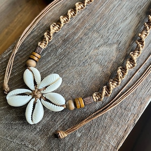 May include: A handmade necklace featuring a flower design made of white seashells. The necklace has a macrame cord in a light brown color, with wooden beads and a sliding knot closure. The necklace is on a wooden surface.