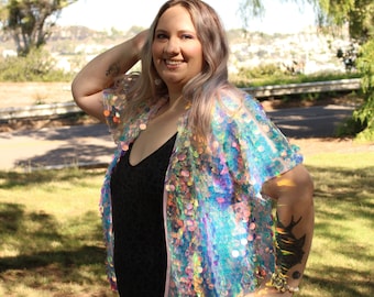 May include: A woman wearing a black swimsuit with a iridescent pink and blue sequined jacket. She is standing in a grassy area with trees in the background.