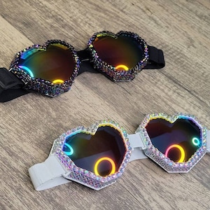 Op de afbeelding: Twee paar hartvormige brillen met iriserende lenzen. Het ene paar heeft een zwart bandje en het andere een wit bandje. Beide paren zijn versierd met strass.