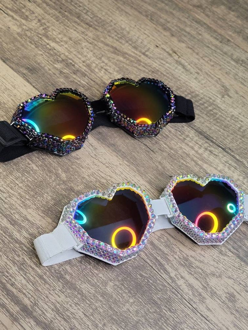 Op de afbeelding: Twee paar hartvormige brillen met iriserende lenzen. Het ene paar heeft een zwart bandje en het andere een wit bandje. Beide paren zijn versierd met strass.