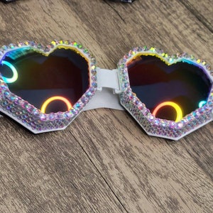 Op de afbeelding: Witte hartvormige bril met regenboogkleurige glazen en een wit elastisch band. De bril is bedekt met strass.