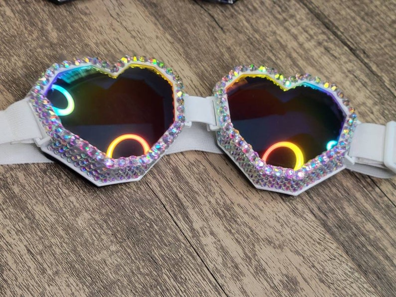 Op de afbeelding: Witte hartvormige bril met regenboogkleurige glazen en een wit elastisch band. De bril is bedekt met strass.