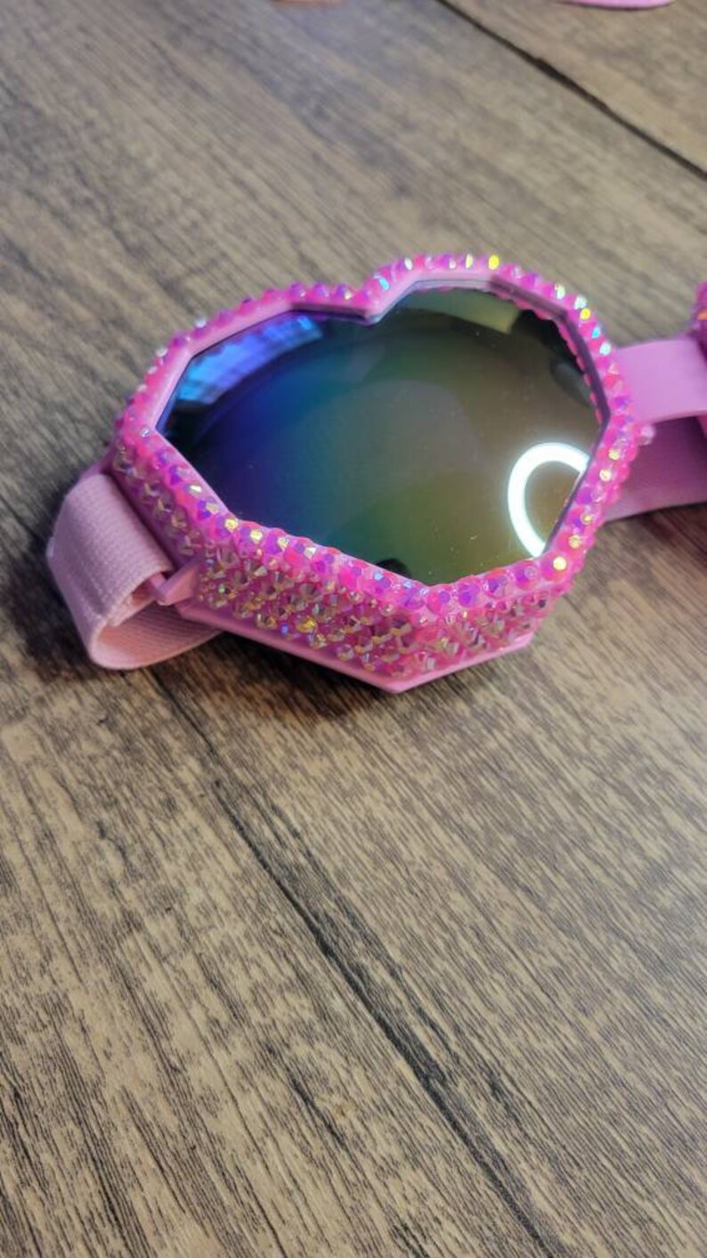 Op de afbeelding: Roze hartvormige zonnebril met iriserende glazen en een roze strassrand.