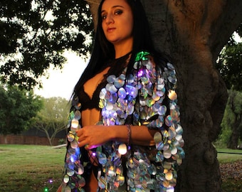 May include: A woman wearing a silver sequined jacket with iridescent accents and a gold crown. The jacket has a black underlay and is decorated with small, round lights. The woman is standing in front of a tree.