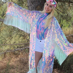 May include: A long, iridescent sequined kimono with a white fringe. The kimono is worn over a pink and blue patterned top and white shorts. The person wearing the kimono is also wearing pink heart-shaped goggles.
