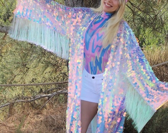 May include: A long, iridescent sequined kimono with a white fringe. The kimono is worn over a pink and blue patterned top and white shorts. The person wearing the kimono is also wearing pink heart-shaped goggles.