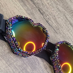 Op de afbeelding: Hartvormige, regenboogkleurige zonnebril met een verstelbare zwarte band. De zonnebril is bedekt met strass en heeft een regenboogkleurige, reflecterende lens.