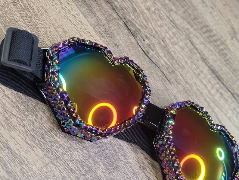 Op de afbeelding: Hartvormige, regenboogkleurige zonnebril met een verstelbare zwarte band. De zonnebril is bedekt met strass en heeft een regenboogkleurige, reflecterende lens.