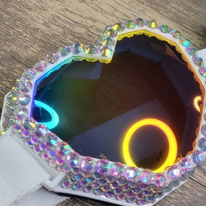 Op de afbeelding: Een wit hartvormig frame met iriserende strasssteentjes rond een regenboogkleurige reflecterende lens. Het frame heeft een witte band.