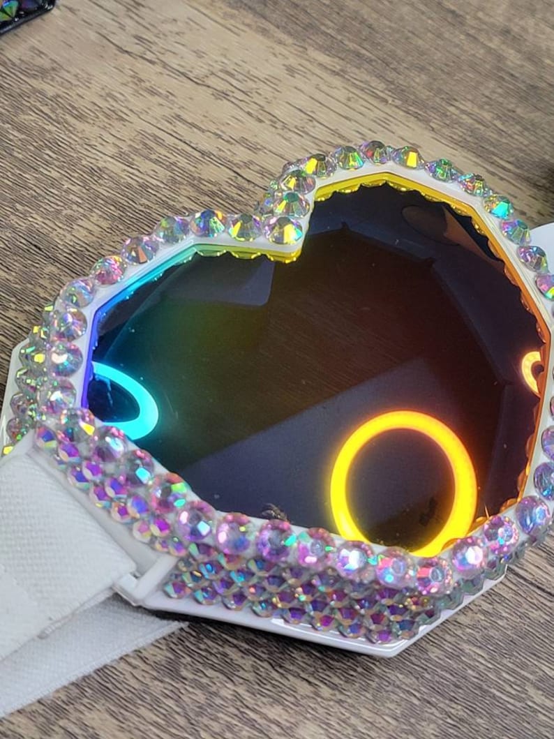 Op de afbeelding: Een wit hartvormig frame met iriserende strasssteentjes rond een regenboogkleurige reflecterende lens. Het frame heeft een witte band.