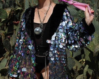 May include: A sequined jacket with a black faux fur collar and iridescent sequins. The jacket is open, revealing a black bodysuit and silver chain accessories. The background features a cactus.
