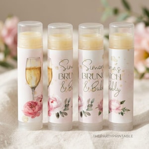 May include: Four lip balm tubes with clear caps. The tubes have a light pink background with floral and champagne glass designs. Text on the tubes reads "Simon's Brunch & Bubbly". The lip balms are a light yellow color.