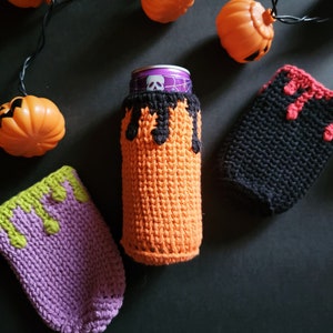 May include: Three crocheted can cozies in orange, purple and black with dripping green, purple and red accents. The cozies are designed to look like Halloween costumes.