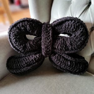 May include: A black knitted bow-shaped pillow. The pillow is made of a soft, textured yarn and has a simple, elegant design.