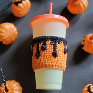 K&ouml;nnte beinhalten: Ein wei&szlig;er To-Go-Becher mit einer schwarzen und orangefarbenen geh&auml;kelten Becherh&uuml;lle. Die H&uuml;lle hat ein tropfendes schwarzes Design. Der Becher hat einen roten Strohhalm und ist von orangefarbenen K&uuml;rbissen umgeben.