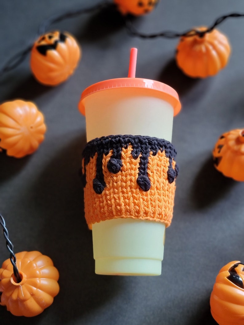 K&ouml;nnte beinhalten: Ein wei&szlig;er To-Go-Becher mit einer schwarzen und orangefarbenen geh&auml;kelten Becherh&uuml;lle. Die H&uuml;lle hat ein tropfendes schwarzes Design. Der Becher hat einen roten Strohhalm und ist von orangefarbenen K&uuml;rbissen umgeben.