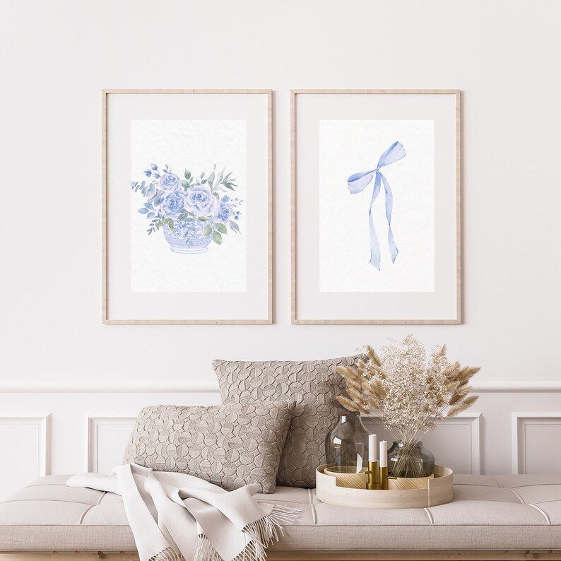Op de afbeelding: Twee ingelijste aquarelprints, een met een blauwe bloemstuk in een vaas en de andere met een blauw lint gebonden in een strik.