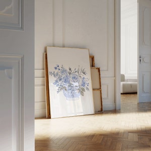 Op de afbeelding: Een aquarel van blauwe rozen in een blauw-witte porseleinen vaas. Het schilderij is ingelijst in een gouden lijst en leunt tegen een witte muur.