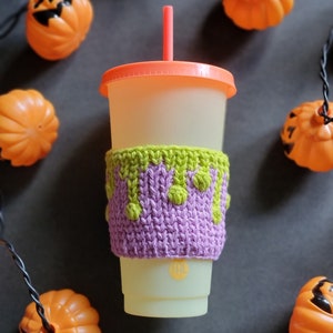 K&ouml;nnte beinhalten: Eine gestrickte Becherh&uuml;lle in Lila und Gr&uuml;n mit einem tropfenden Schleim-Design. Die H&uuml;lle befindet sich auf einem wei&szlig;en To-Go-Becher mit rosafarbenem Deckel und rotem Strohhalm. Der Becher ist von orangefarbenen K&uuml;rbissen mit Jack-o'-Lantern-Gesichtern umgeben.