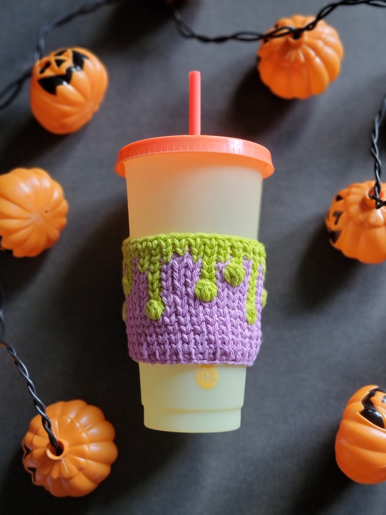 K&ouml;nnte beinhalten: Eine gestrickte Becherh&uuml;lle in Lila und Gr&uuml;n mit einem tropfenden Schleim-Design. Die H&uuml;lle befindet sich auf einem wei&szlig;en To-Go-Becher mit rosafarbenem Deckel und rotem Strohhalm. Der Becher ist von orangefarbenen K&uuml;rbissen mit Jack-o'-Lantern-Gesichtern umgeben.