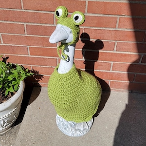 May include: A white goose statue wearing a lime green crocheted frog hat and sweater. The hat has large, round eyes. The goose is standing on a decorative base, with a brick wall in the background.