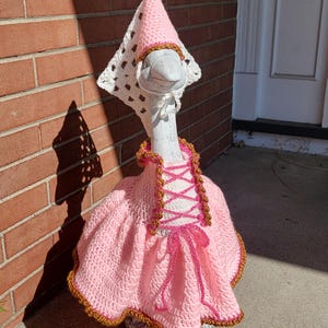 Puede incluir: Una estatua decorativa de ganso con un vestido y un gorro de ganchillo rosa hechos a mano. El vestido presenta un frente estilo corsé con lazos rosas y ribete dorado. El gorro es blanco con la parte superior rosa y ribete dorado.