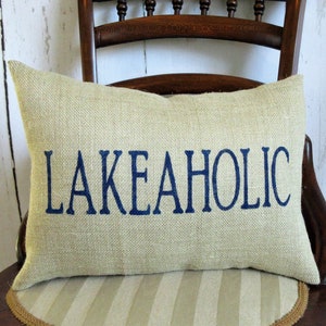 May include: A beige burlap pillow with the text "LAKEAHOLIC" embroidered in navy blue.