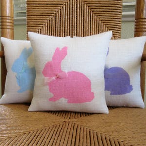 May include: Three decorative throw pillows with white fabric and bunny silhouettes. The bunnies are pink, blue, and purple. The pillows are sitting on a wicker chair.