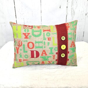 May include: Rectangular burlap pillow with a colorful letter design in green, orange, and red. A red band with three yellow buttons accents the right side. The pillow is on a white surface.