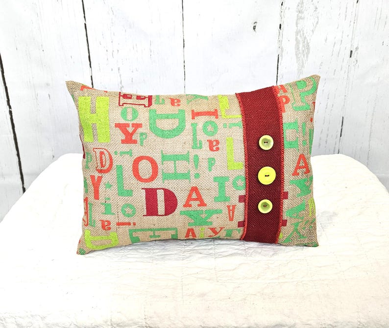 May include: Rectangular burlap pillow with a colorful letter design in green, orange, and red. A red band with three yellow buttons accents the right side. The pillow is on a white surface.