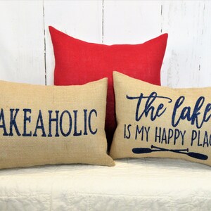 May include: Three decorative throw pillows with burlap fabric and navy blue lettering. The pillows feature the phrases "Lake Aholic", "The Lake is My Happy Place" and a pair of oars.