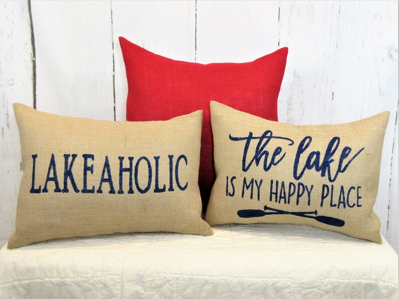 May include: Three decorative throw pillows with burlap fabric and navy blue lettering. The pillows feature the phrases "Lake Aholic", "The Lake is My Happy Place" and a pair of oars.
